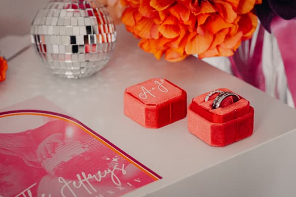 Close-up of two wedding rings in a red velvet ring box with initials 'A & J' on the lid, placed on a white surface next to an orange flower bouquet and a small disco ball at Sandstone Point Hotel.