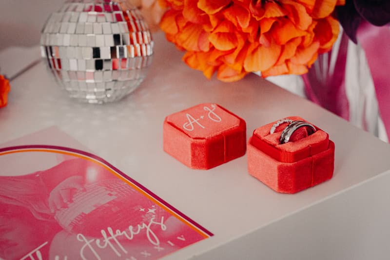 Close-up of two wedding rings in a red velvet ring box with initials 'A & J' on the lid, placed on a white surface next to an orange flower bouquet and a small disco ball at Sandstone Point Hotel.