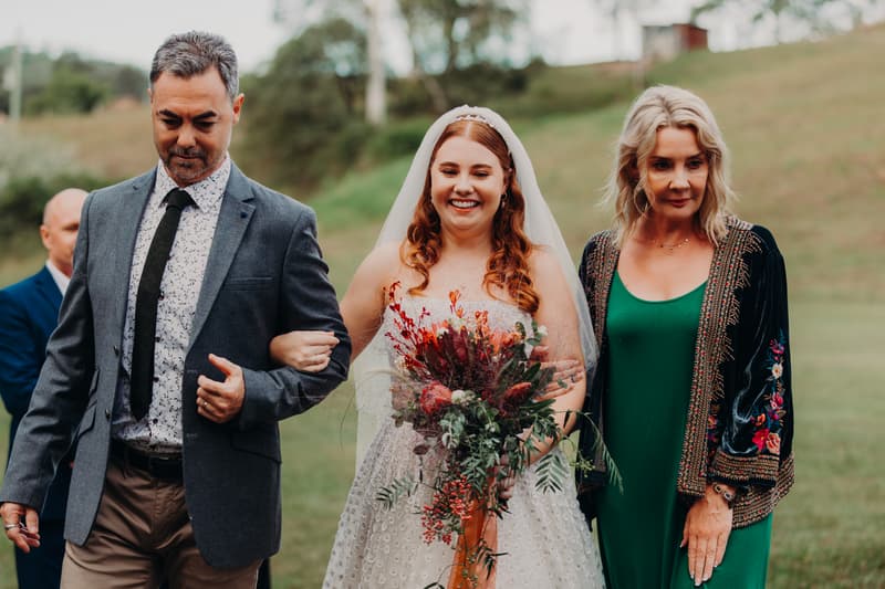 The bride Lilly, holding a bouquet, walks arm-in-arm with an older man and an older woman outdoors at Yabbaloumba Retreat — By The River.