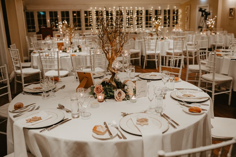 Round wedding reception tables set with white tablecloths, white plates, cutlery, glasses, bread rolls, and floral centerpieces with decorative branches and hanging glass candle holders. White chairs surround the tables and fairy lights decorate the background.