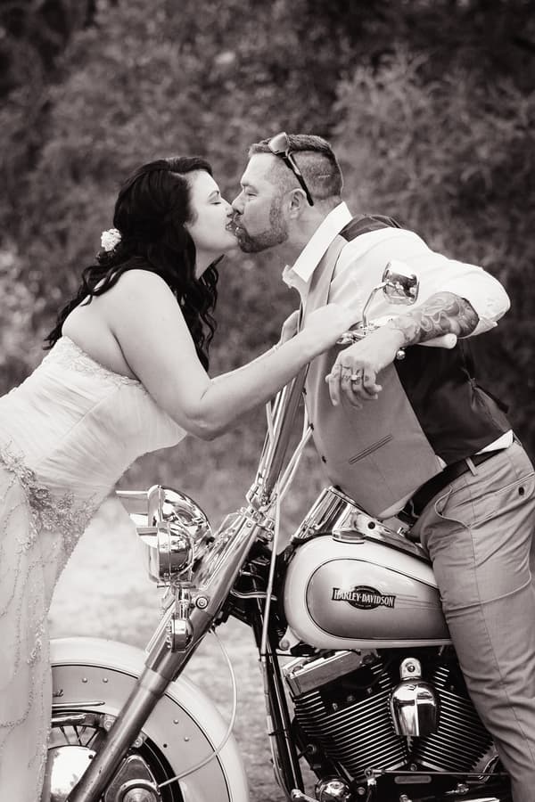Bride Kelly and groom Shane kiss while Shane leans on a Harley Davidson motorcycle outdoors.
