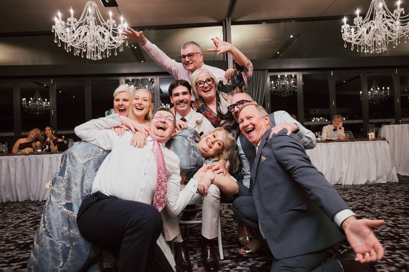 Joel and Morgan pose with a group of guests at the reception in The Pumicestone Room, Sandstone Point Hotel, under chandeliers with tables and seated guests in the background.