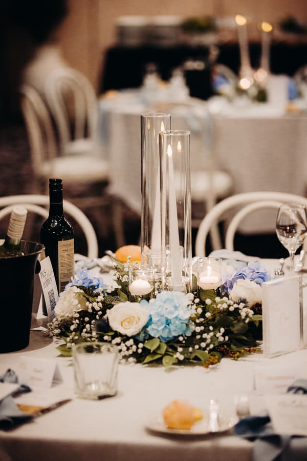 Table centerpiece at the Sandstone Point Hotel Pumicestone Room reception featuring white and blue floral arrangement with candles in glass holders, wine bottles, and place settings.