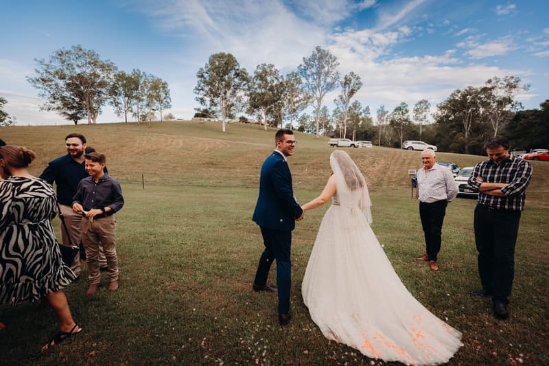 Bride Lilly and groom Connor hold hands on a grassy field at Yabbaloumba Retreat — By The River, surrounded by guests standing and chatting under a partly cloudy sky.