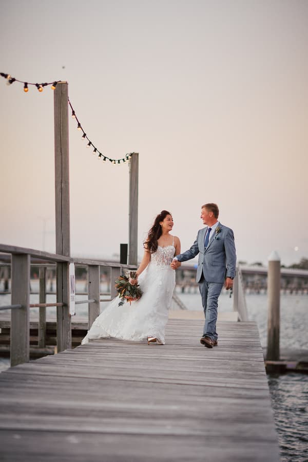 Bride Jheng and groom Dane walk hand in hand on a wooden pier at Sandstone Point Hotel, with Jheng holding a bouquet and both smiling at each other.