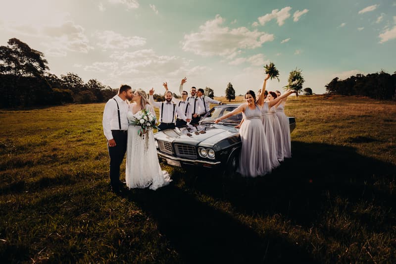 Bride Annie and groom Trent kiss beside a vintage car outdoors at Ocean View Estates, surrounded by bridesmaids in lavender dresses holding bouquets and groomsmen in white shirts with suspenders.