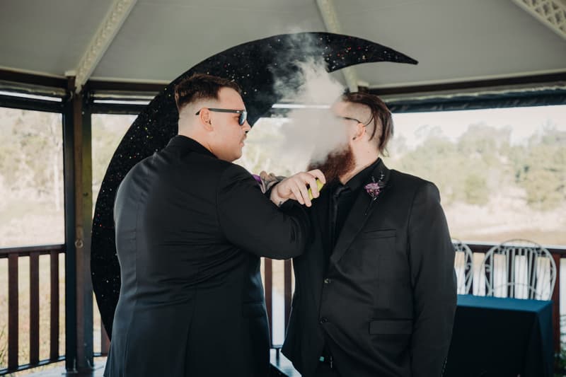 Two men in black suits stand on the ceremony stage at Ocean View Estates — On The Lake, with one man exhaling vapor from a vape device held by the other.