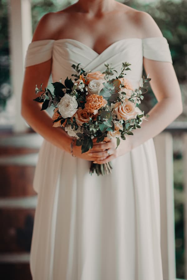 The bride Courtney in an off-shoulder white wedding dress holds a bouquet of mixed flowers including roses and greenery at Tiffany's Maleny.