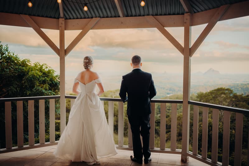 The bride Courtney and groom Liam stand side by side on a covered balcony at Tiffany's Maleny, looking out over a scenic landscape with distant mountains under a partly cloudy sky.