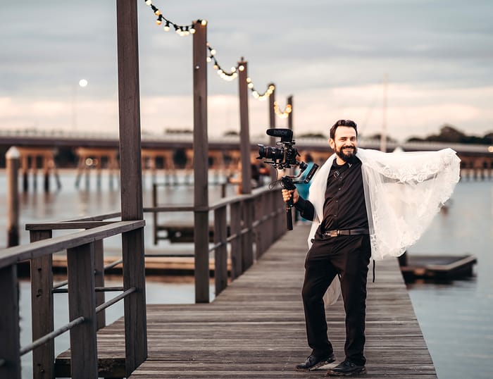 Roman at the Sandstone Point Jetty wearing a black outfit and the bride's veil, smiling and holding a small cinema camera on a gimbal
