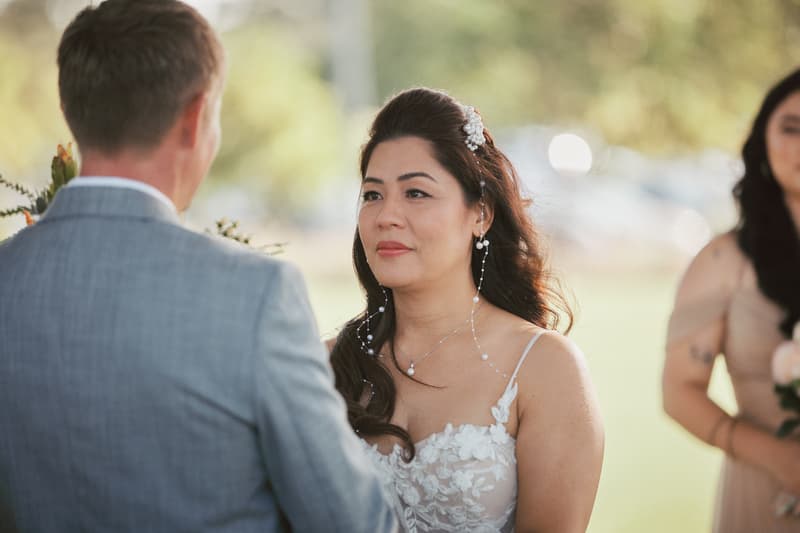 Bride Jheng and groom Dane face each other during their wedding ceremony at Sandstone Point Hotel — The Pavilion, with a bridesmaid partially visible in the background.