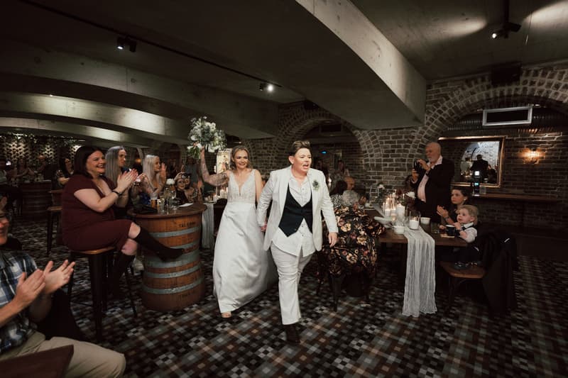 Brides Mandy and Selina walk hand in hand through the reception area at Sandstone Point Hotel — The Cellar, with Mandy holding a bouquet and guests clapping and taking photos.