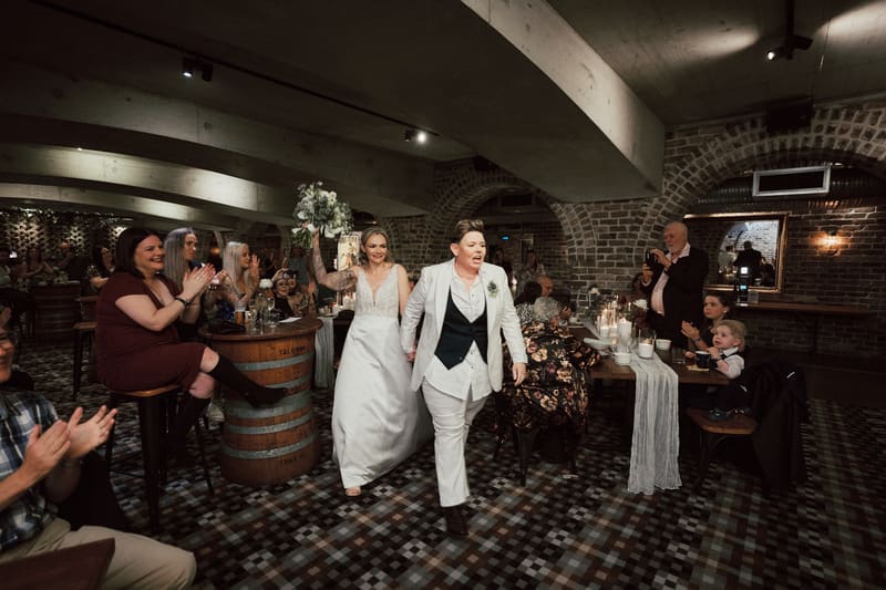Brides Mandy and Selina walk hand in hand through the reception area, with Mandy holding a bouquet and guests clapping and taking photos. Winter wedding light photography Brisbane by She Said Yes.