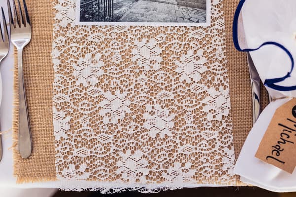 Close-up of a wedding reception table setting at TOOWONG ROWING CLUB — The Malouf Room, featuring a lace table runner over a burlap placemat, silver cutlery, a white napkin with blue trim, and a name tag labeled 'Michael'.