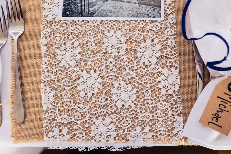 Close-up of a wedding reception table setting at TOOWONG ROWING CLUB — The Malouf Room, featuring a lace table runner over a burlap placemat, silver cutlery, a white napkin with blue trim, and a name tag labeled 'Michael'.