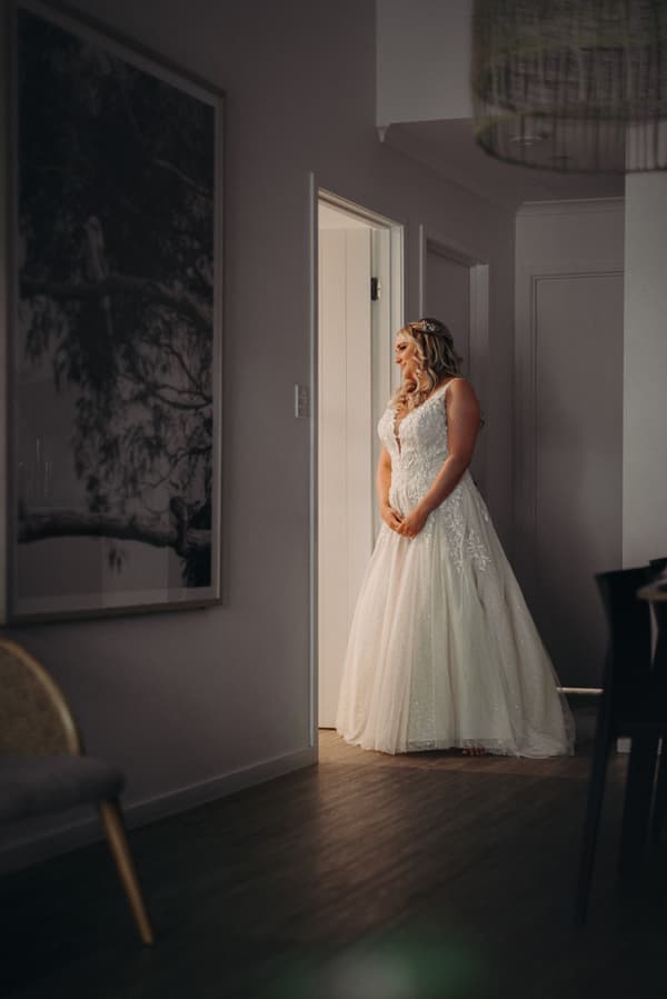 The bride Chloe stands alone in a hallway at Sandstone Point Hotel, wearing a white lace wedding gown and looking out through a doorway.