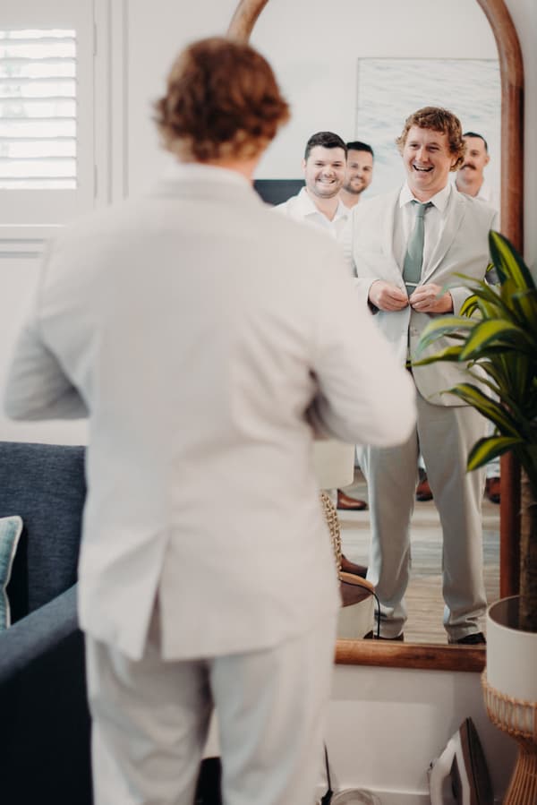 Brodie adjusts his light-colored suit jacket while looking into a mirror, with three groomsmen smiling behind him at Sandstone Point Hotel.
