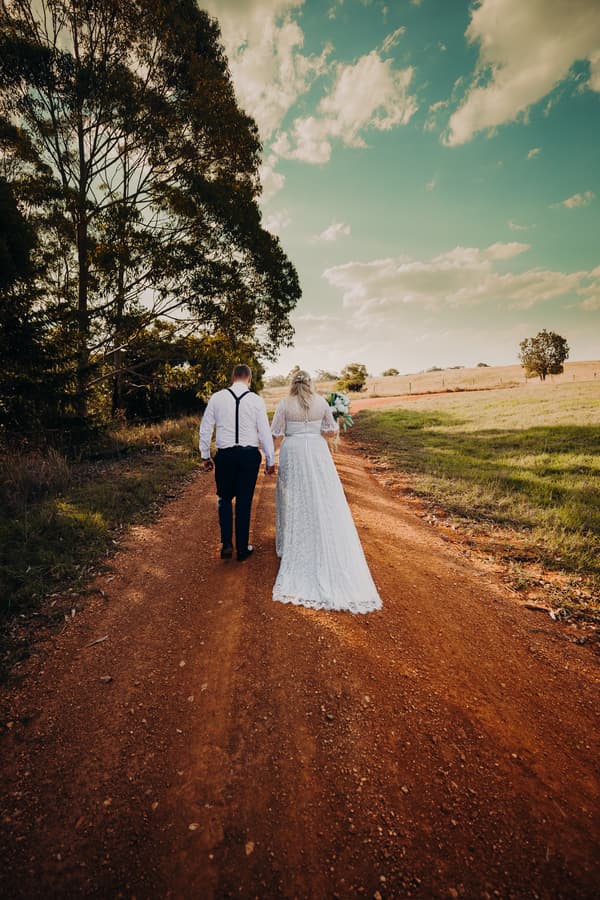 Bride Annie and groom Trent walk hand in hand down a dirt path at Ocean View Estates, with trees on one side and open fields on the other under a partly cloudy sky.