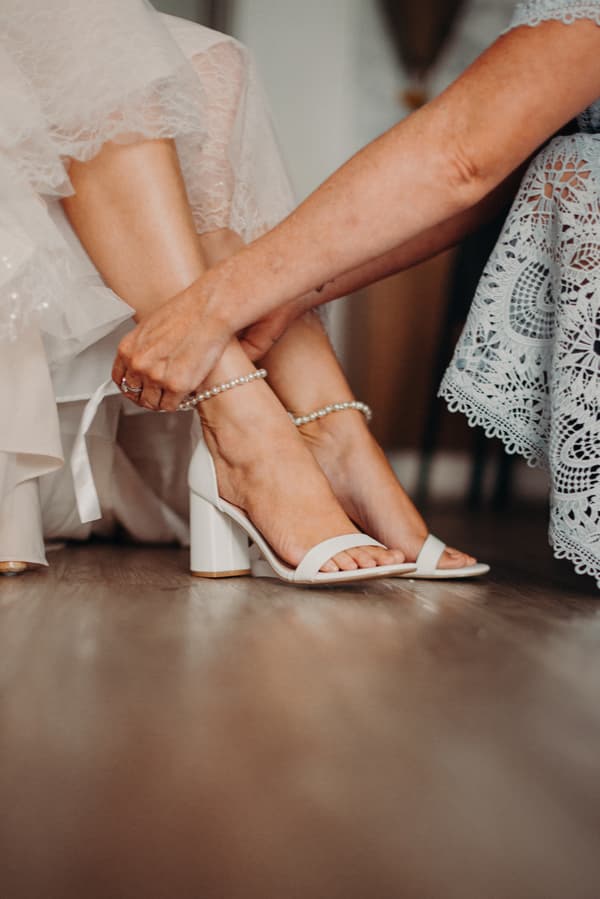 The mother of the bride fastens a pearl anklet on the bride's foot as the bride wears white heeled sandals and a lace wedding dress at Sandstone Point Hotel.