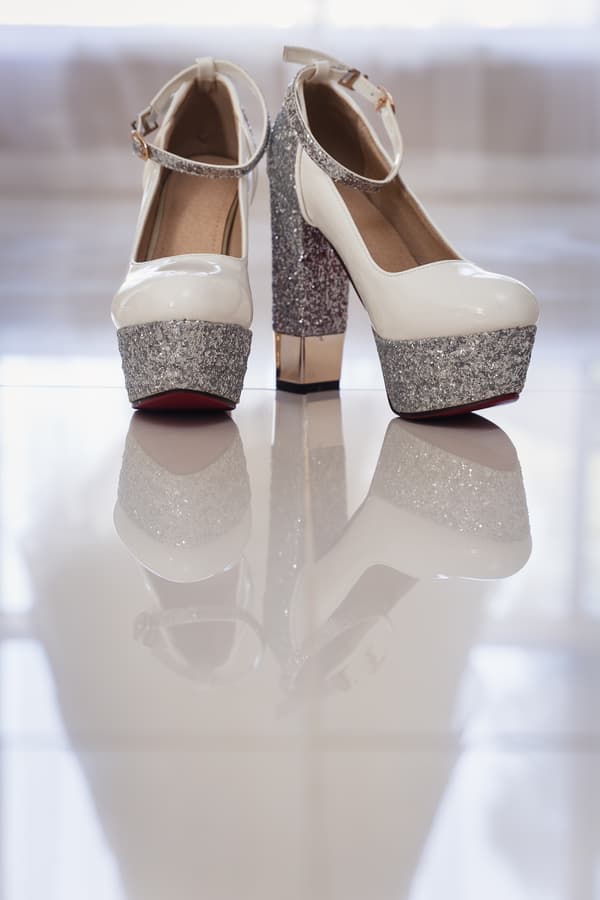 Close-up of the bride's white high-heeled shoes with silver glitter platform and ankle straps, reflected on a shiny floor at Kwila Lodge.
