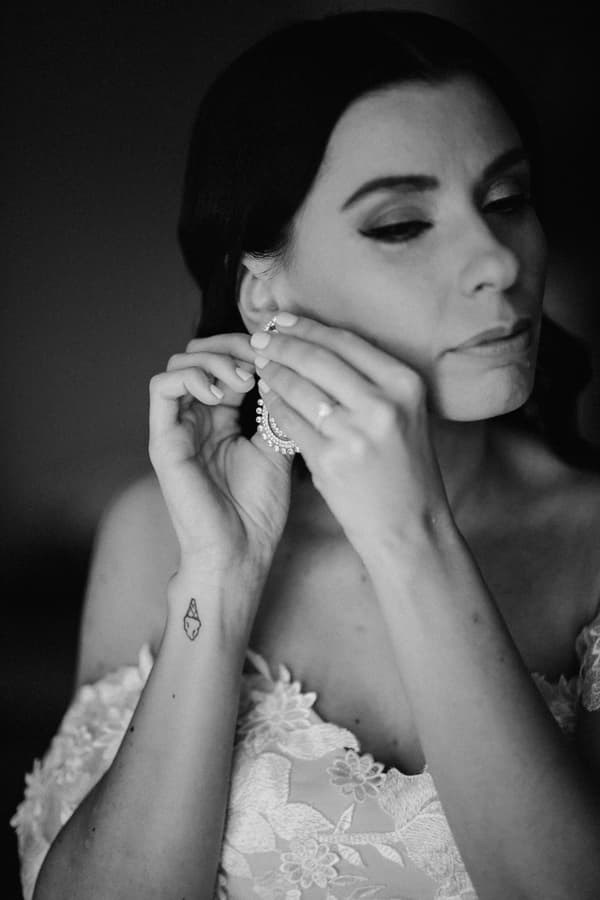 Bride in a lace off-shoulder wedding dress putting on a large hoop earring, shown in a close-up black and white image.