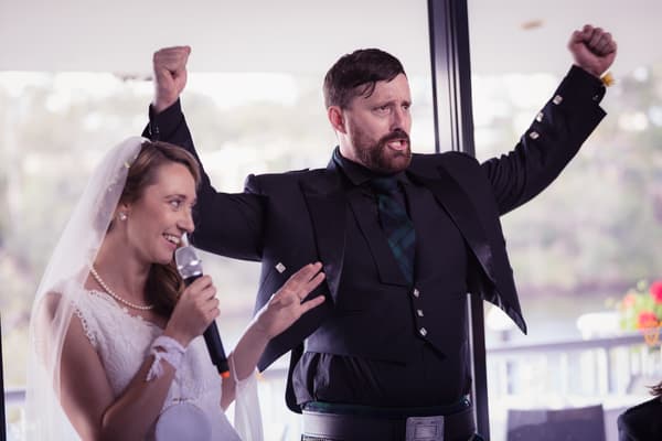The bride Francesca holds a microphone and speaks while the groom Ben raises both arms in celebration at the reception in The Malouf Room, Toowong Rowing Club.