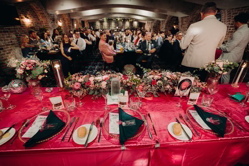 Reception guests seated at long tables inside Sandstone Point Hotel — Cellar, with the head table in the foreground decorated with pink floral arrangements, menus, and place settings labeled for Mindy and David.