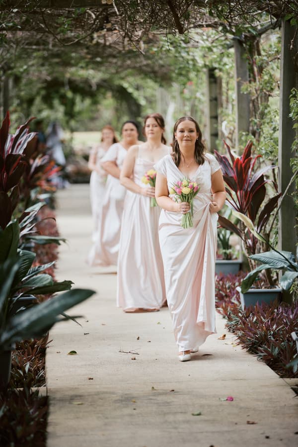 Four bridesmaids in pale pink dresses walk in a line holding bouquets along a garden pathway at Eatons Hill Hotel — Lakeside.