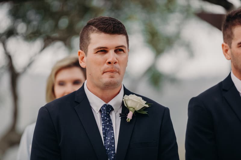 The groom Brent stands at the ceremony stage at Preston Peak Winery Functions Centre wearing a dark suit with a white rose boutonniere and a patterned tie, with a blurred woman visible behind him.