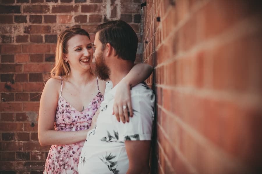 The couple leaning against a brick wall, smiling warmly at each other at Brisbane Powerhouse.