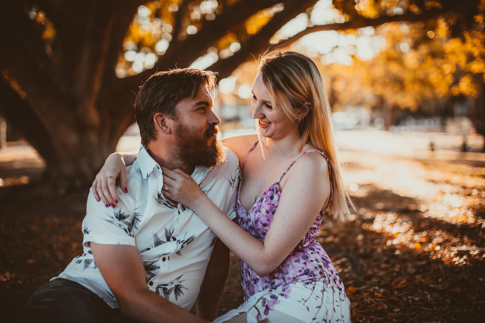 The couple sitting on a picnic blanket under a large tree, with sunlight filtering through the leaves.