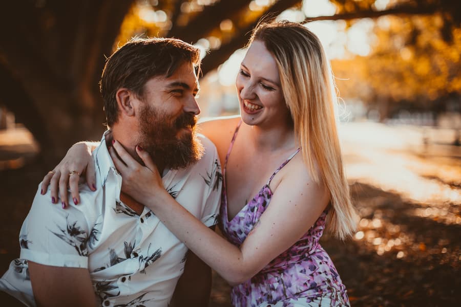 The couple sitting close on a blanket under a tree at Brisbane Powerhouse, smiling warmly at each other in soft, golden light.