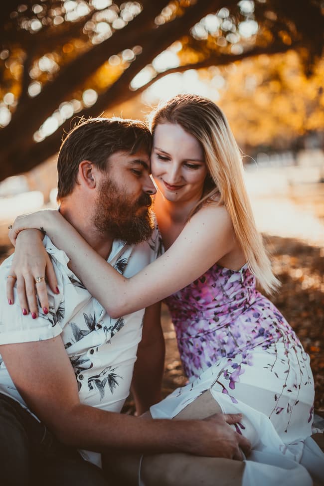 The couple sitting on the ground, embracing affectionately under a tree at Brisbane Powerhouse, with a warm, intimate expression.