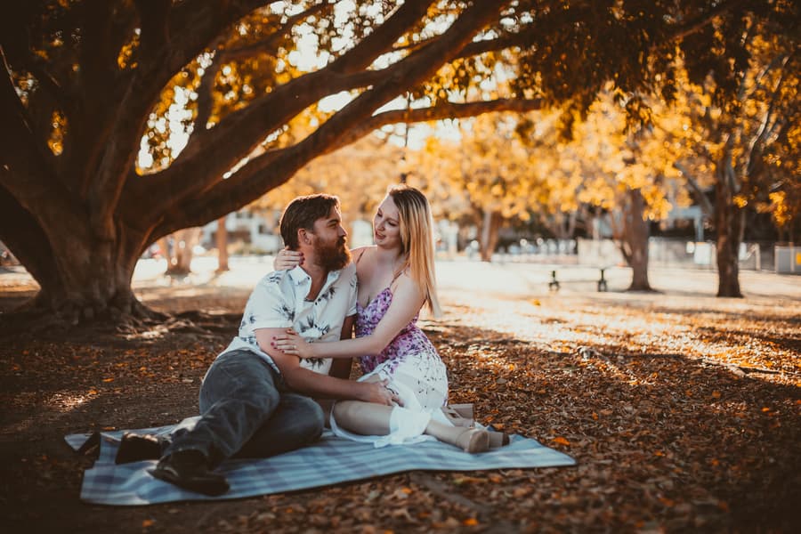 The couple sitting on a blanket under a large tree at Brisbane Powerhouse, surrounded by fallen leaves, sharing a tender moment.