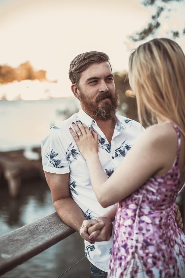 The couple standing on a balcony at Brisbane Powerhouse, with one resting a hand on the other's chest, looking into each other's eyes.