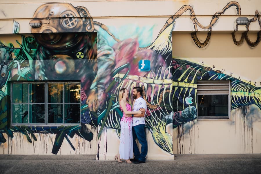 The couple standing in front of a vibrant mural at Brisbane Powerhouse, holding each other close and smiling.
