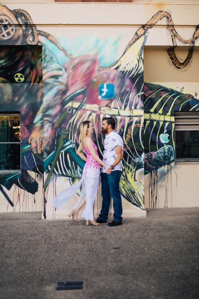 The couple standing in front of a mural at Brisbane Powerhouse, holding hands and looking at each other with joy.