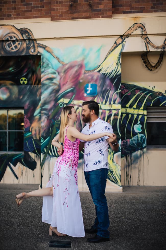 The couple dancing playfully in front of a mural at Brisbane Powerhouse, with one lifting a leg in a playful pose.