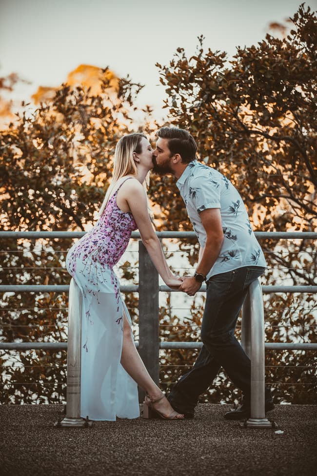 The couple leaning over a railing at Brisbane Powerhouse, sharing a kiss with trees in the background.
