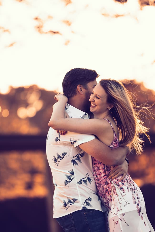 The couple embracing warmly, smiling under a sunset sky at Brisbane Powerhouse.