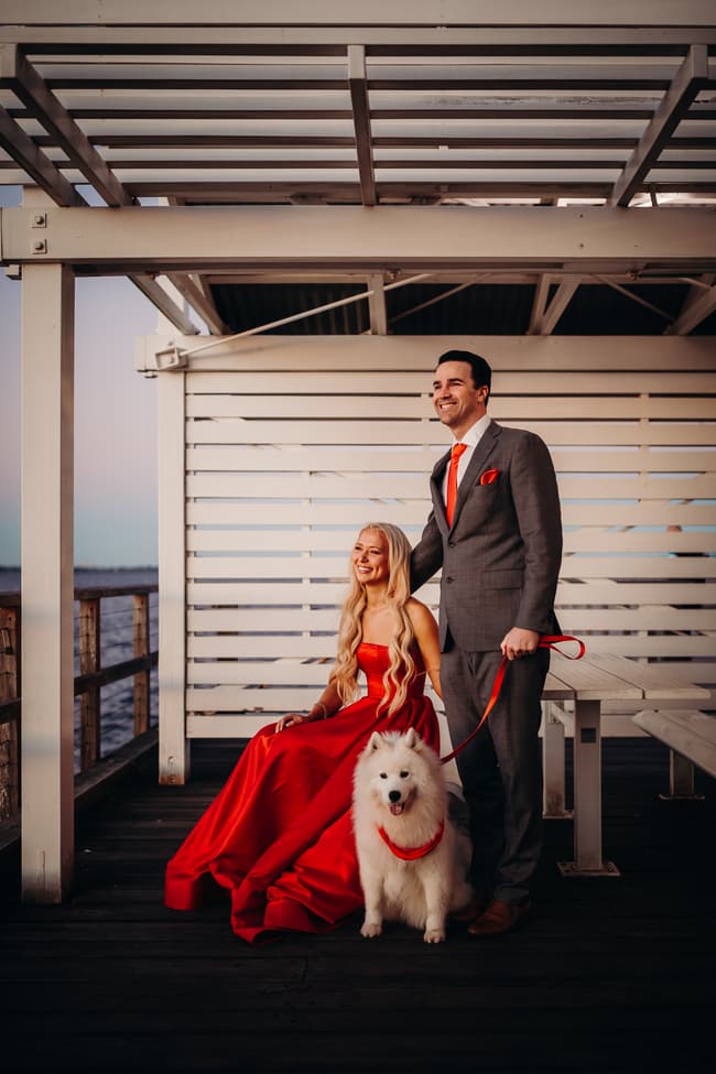 The couple sits on a bench at Woody Point Jetty with a fluffy white dog. The setting sun casts a warm glow.