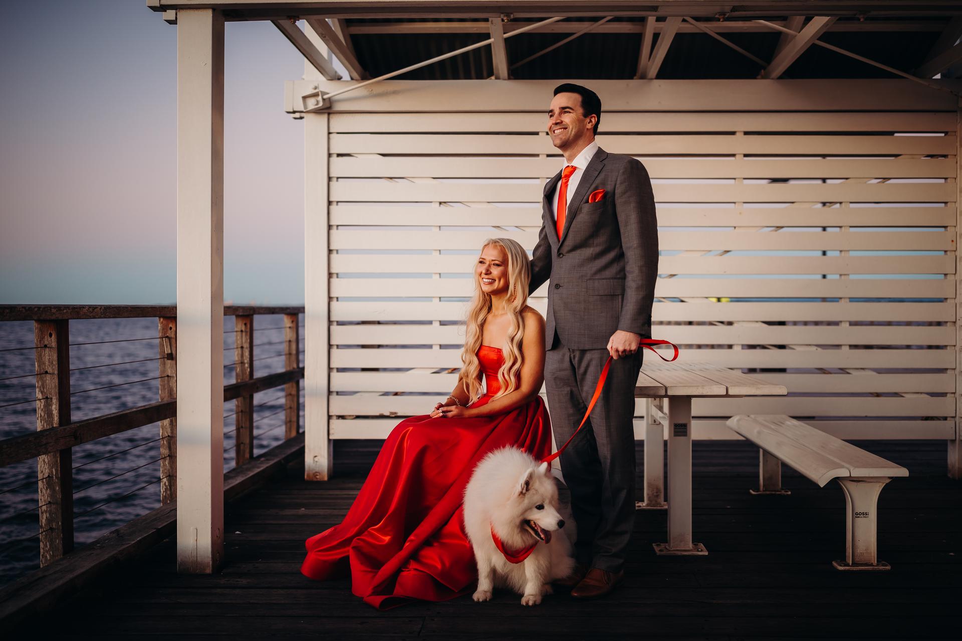 The couple stands on a bench at Woody Point Jetty, smiling with their dog. The sky is a soft pastel hue.
