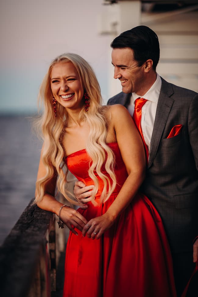 The couple leans against the railing at Woody Point Jetty, laughing together as the sun sets.
