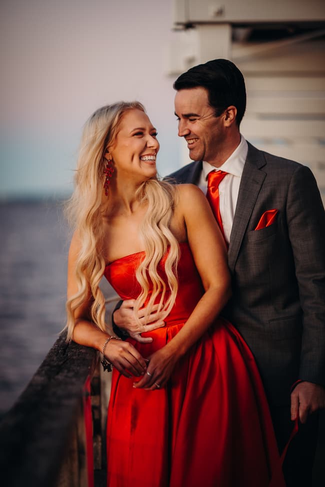 The couple stands close, smiling at each other on Woody Point Jetty. The ocean is visible in the background.