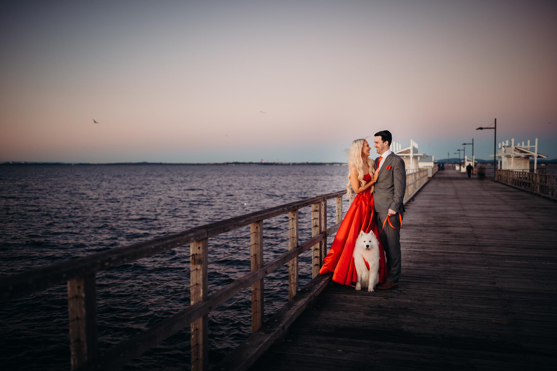 The couple stands on the jetty, gazing into each other's eyes with the ocean behind them.