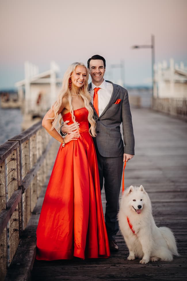The couple stands on the jetty with their dog at sunset, the sky painted in soft colors.