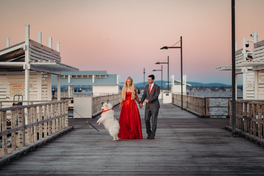 The couple poses on the jetty with their dog, both smiling warmly at the camera.