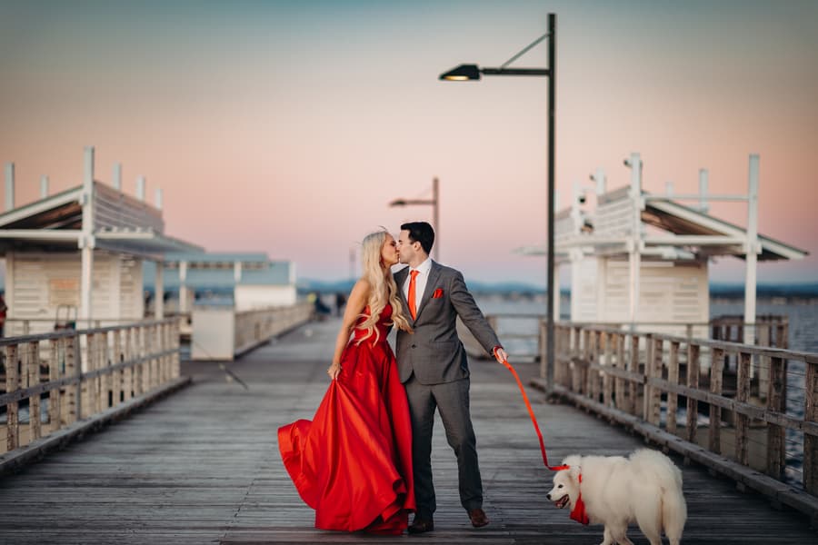 The couple walks hand in hand on the jetty with their dog, the sky a gentle pink.
