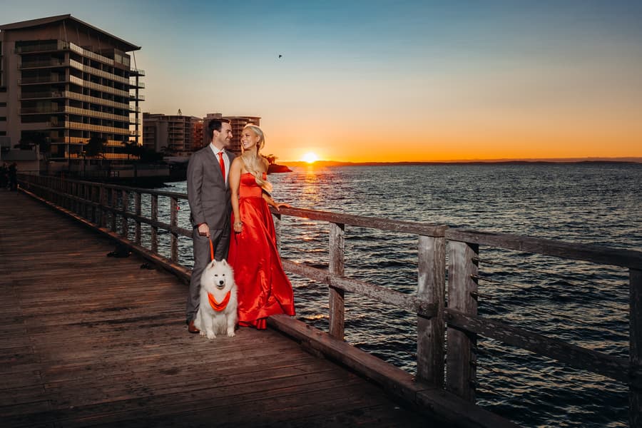The couple shares a kiss on the jetty while their dog looks on. The sky is a soft pastel.