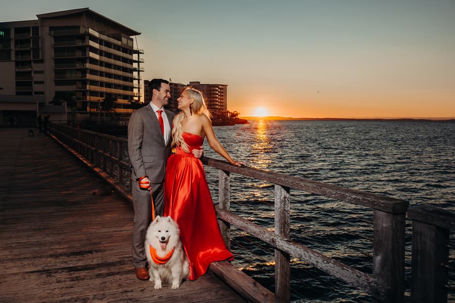The couple stands by the railing at sunset, the ocean and sky glowing warmly.